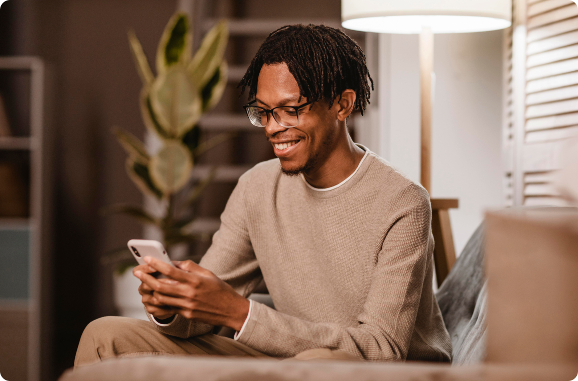 Homem negro usando óculos, sentado no sofá, feliz e com um celular na mão